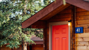Tan exterior of guest cabin #41 at Denali Cabins with red door & porch light in a forested setting on a sunny day in Alaska.