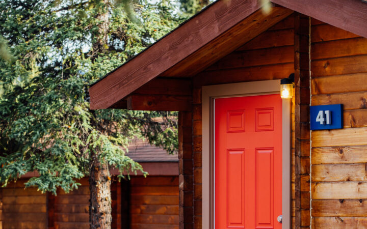 Tan exterior of guest cabin #41 at Denali Cabins with red door & porch light in a forested setting on a sunny day in Alaska.