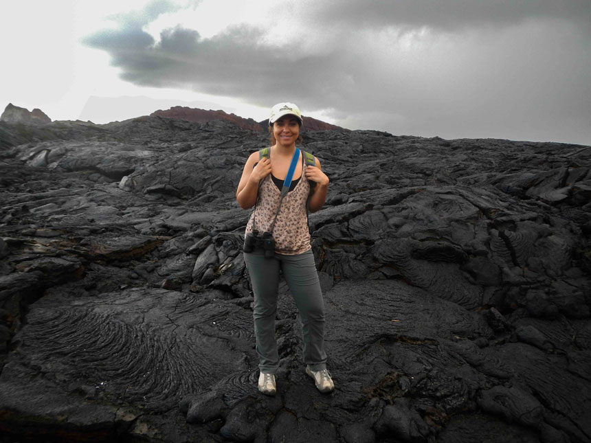 Woman in beige tank top & gray pants stands atop cooled black lava on a cloudy day in the Galapagos Islands.