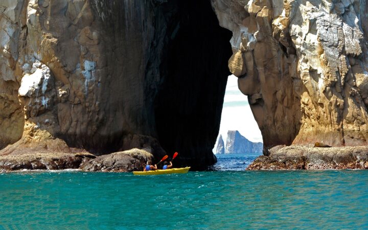2 travelers paddle a yellow double kayak through the arch of a giant rock structure in the Galapagos, in the distance another iconic rock landmark, Kicker Rock.