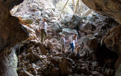 Man, woman & 2 kids walk down into a cave over large beige rocks under a sunny sky during a Galapagos Safari land tour.