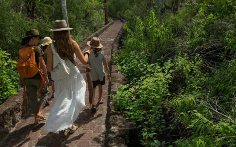 Woman in felt hat & white sundress walks with 3 kids on a stone pathway among bright green jungle during a Galapagos Safari.
