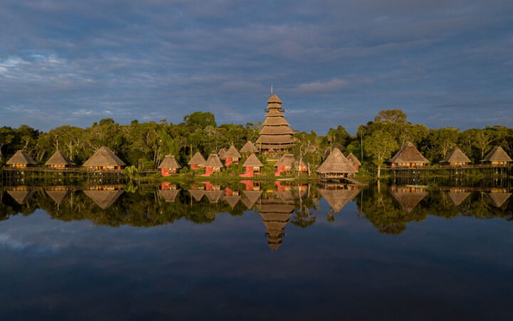 Napo Wildlife Center in Ecuador's Amazon with thatched-roof triangular buildings sitting on glassy lake with jungle behind at dusk.