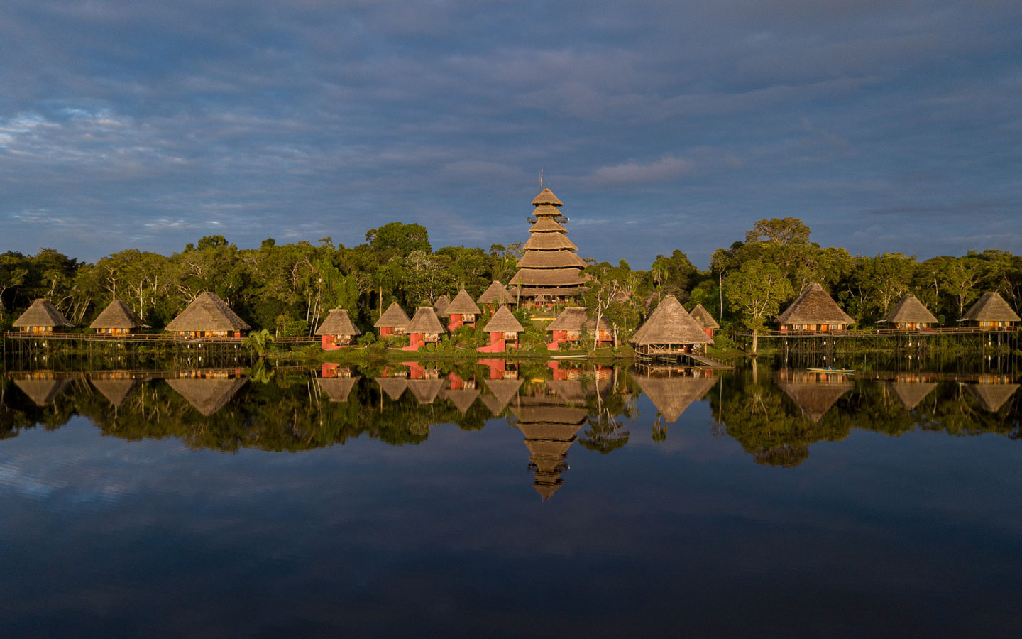 Napo Wildlife Center in Ecuador's Amazon with thatched-roof triangular buildings sitting on glassy lake with jungle behind at dusk.