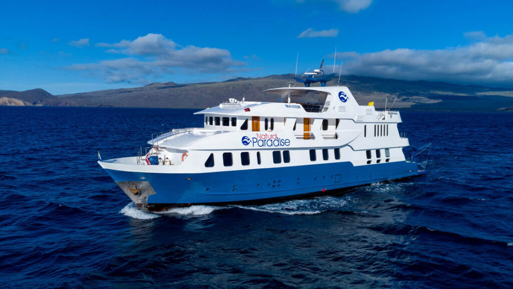 Exterior port side of Natural Paradise Galapagos small ship cruising with blue hull & 3 white upper decks.