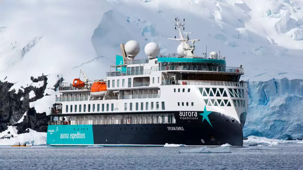 Sylvia Earle ship sitting in calm water, with blue & teal exterior with star logo, 5 white upper decks & 2 orange life boats.