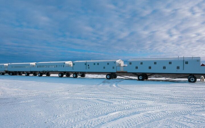 The Tundra Lodge on wheels on a flat snow covered landscape in the Canadian Arctic on a cloudy day with one polar bear nearby.