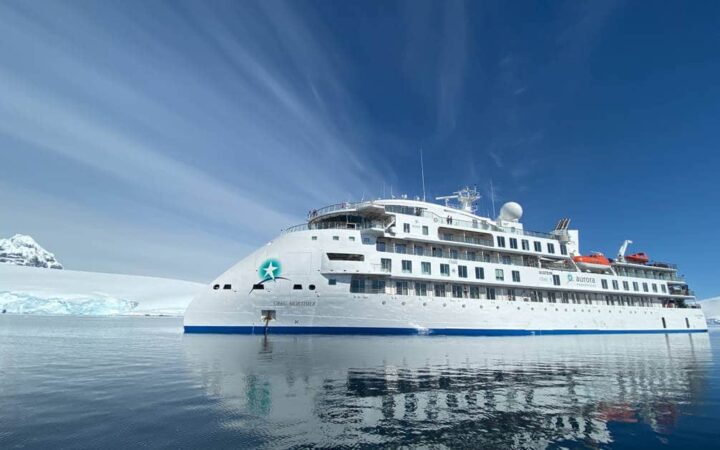 Exterior of Greg Mortimer polar expedition ship, with pointed bow, white exterior & 6 passenger decks, on a sunny day in Antarctica.