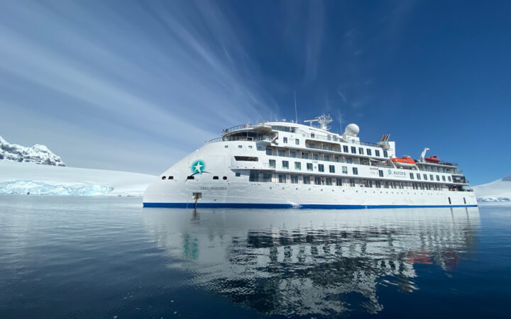 Exterior of Greg Mortimer polar expedition ship, with pointed bow, white exterior & 6 passenger decks, on a sunny day in Antarctica.