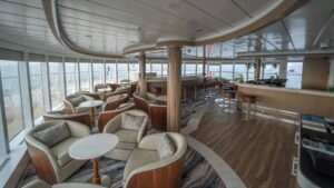 Observation Deck Bar aboard Greg Mortimer polar ship, with white-&-wood lounge chairs arranged around small marble-&-wood tables, surrounded by picture windows.
