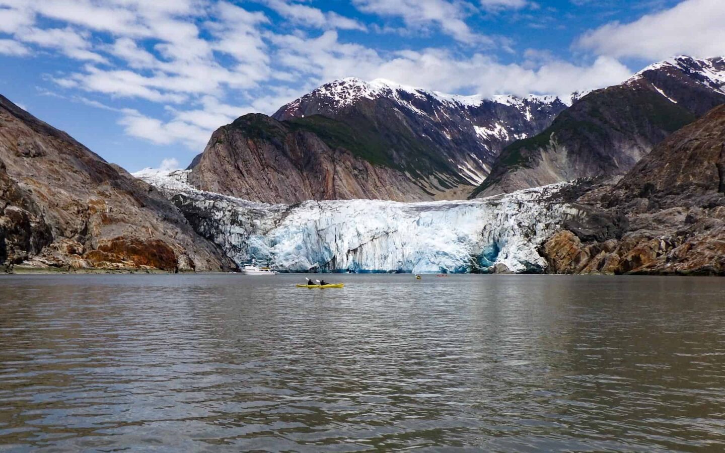 Alaska travelers with guide kayaking in front of a glacier and a small ship in the background.