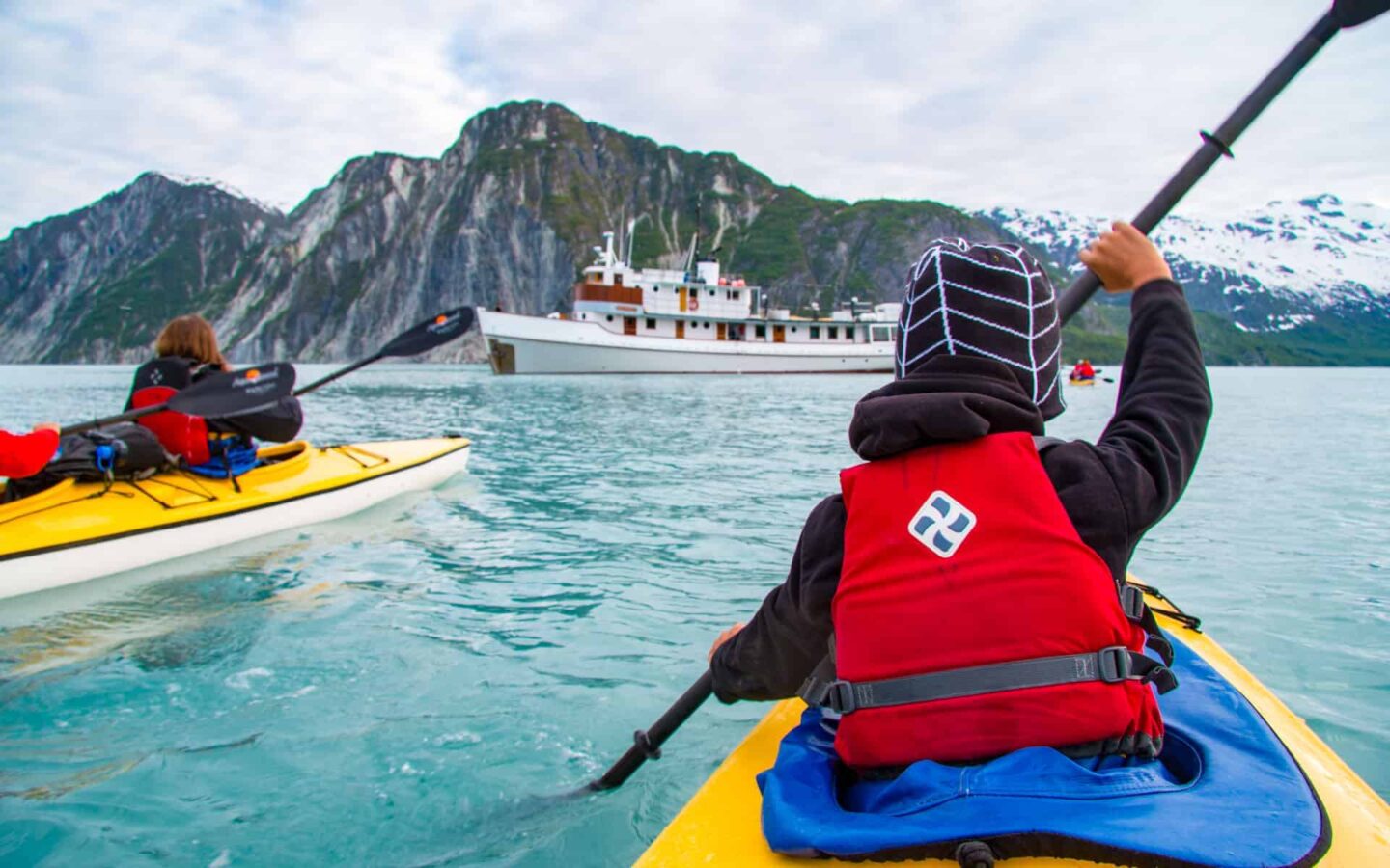 two adventure travelers in yellow kayaks paddling toward a small ship in bright blue glacial water on an alaska trip