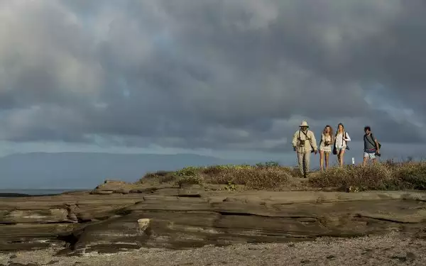 Three travelers with cameras hike with their Galapagos guide on rocks with a cloudy sky