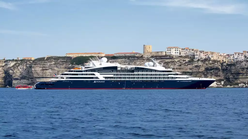 Le Laperouse small expedition ship with dark blue hull & white upper decks sits in calm blue sea beside cliffside town in sun.