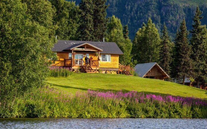The Winterlake Lodge is perched on a grassy knoll above flowers and water, with pine trees in the background, in Alaska