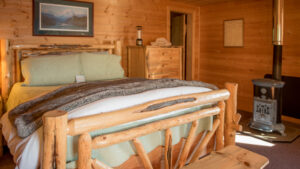 Wood post & beam double bed with sea green sheets & white comforter by small gas fireplace in a rustic wooden cabin.
