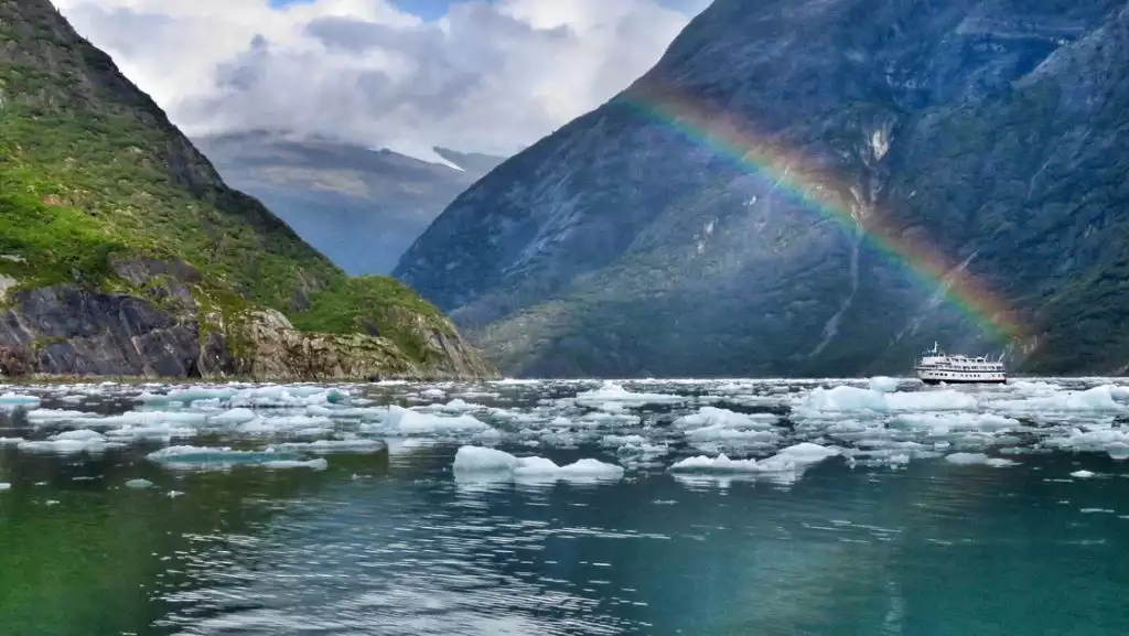 Small ship with blue exterior & teal accents sits among iceberg bits under a rainbow between two rocky cliff faces in Alaska.