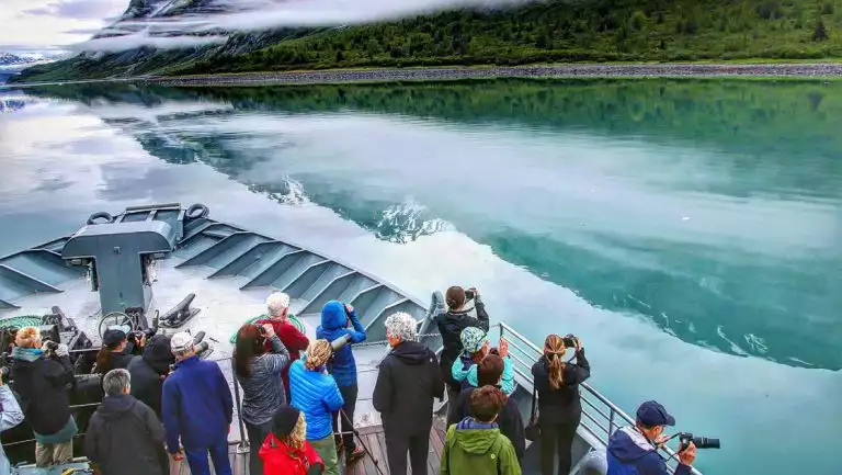 Guests on Alaska's Ultimate Adventure Cruise stand on bow of small ship watching forested shoreline with binoculars & cameras.