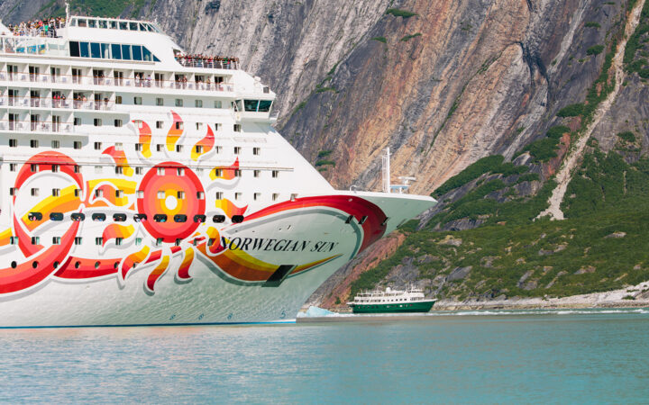 The bow of a big cruise ship dwarfs a small ship in size as they are seen side by side by the rocky wall of a fjord