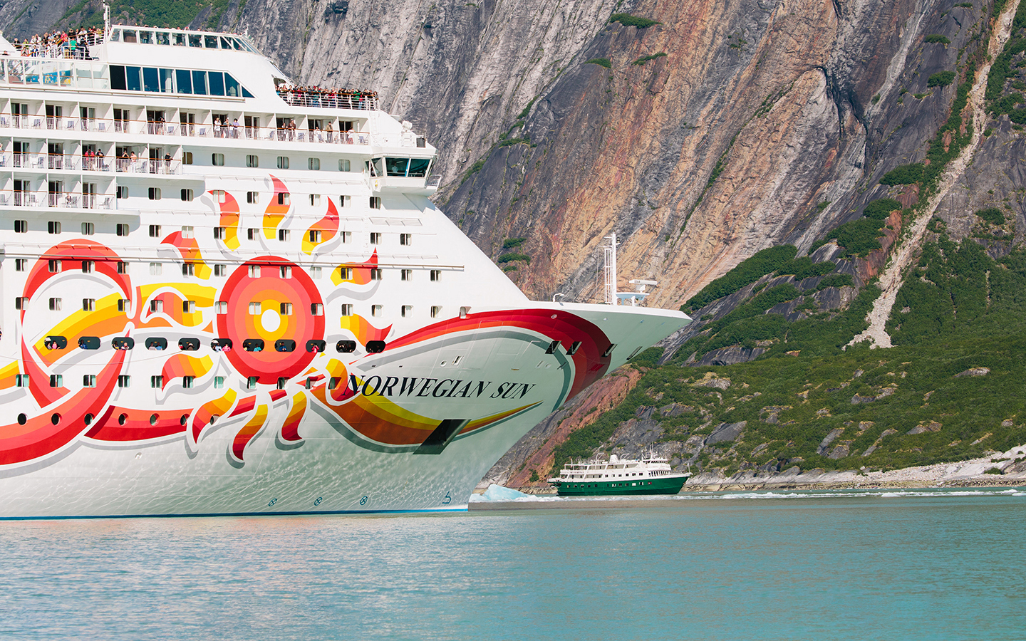 The bow of a big cruise ship dwarfs a small ship in size as they are seen side by side by the rocky wall of a fjord