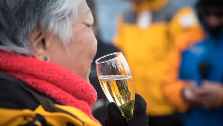 Woman in yellow parka & red neck gaiter holds glass of champagne while toasting crossing the Antarctic Circle on an expedition.