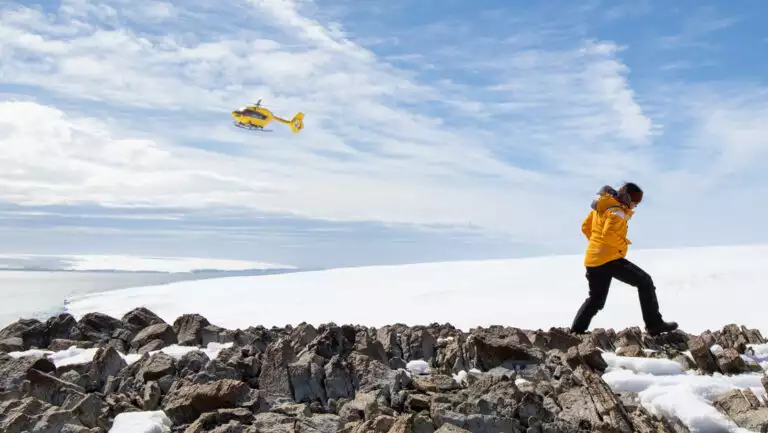 Antarctic Circle traveler in yellow parka walks over rocky peak with snowfield beyond & yellow helicopter flying overhead.