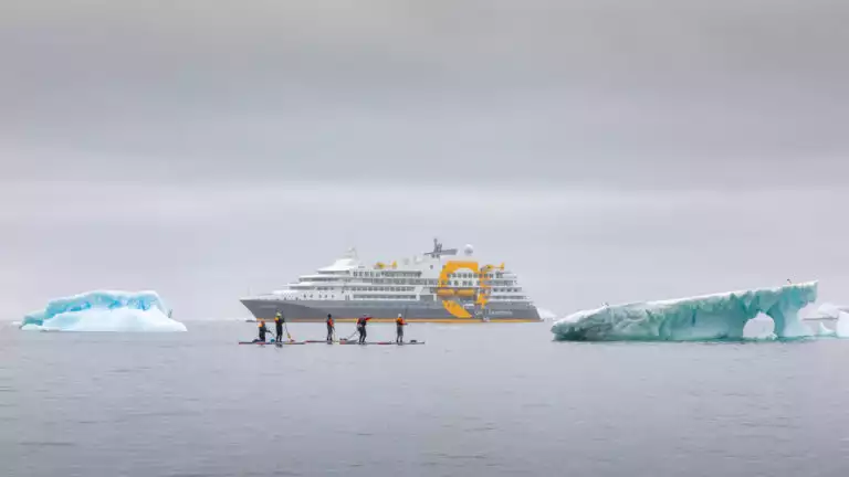 paddle boarders braving the icy cold waters next to two large icebergs in front of Quark expedition cruise ship
