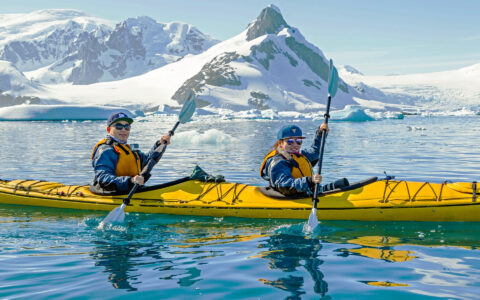 Luxury expedition travelers kayak among icebergs and snowy Antarctic peaks.
