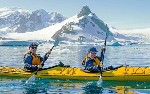 Kayakers paddle a yellow tandem kayak through icy blue waters with snowcapped peaks in the background on a sunny day in Antarctica.