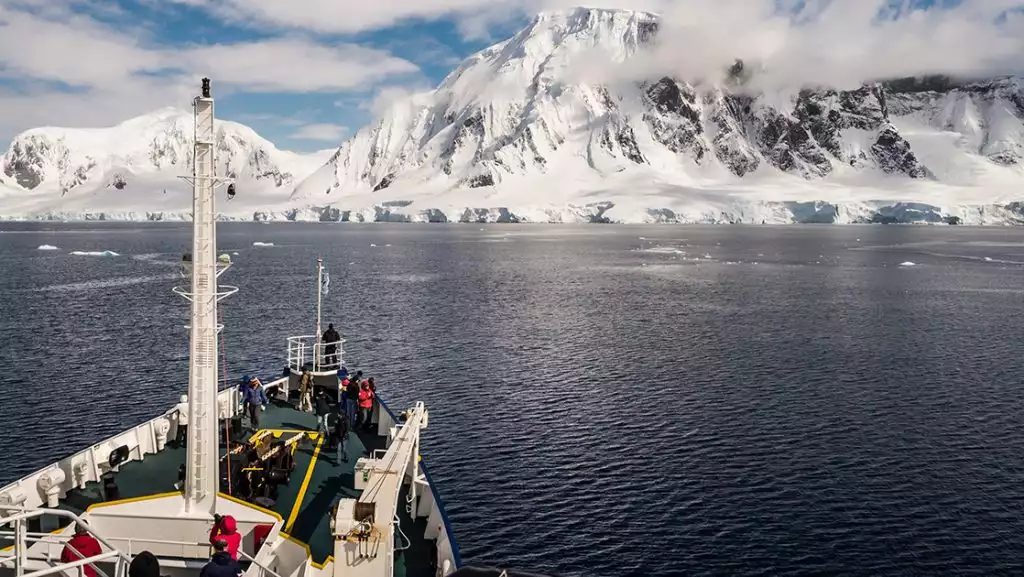 Polar Circle Cruise guests stand on bow of small green & white expedition ship look out at tall, snowy peaks in the sun.