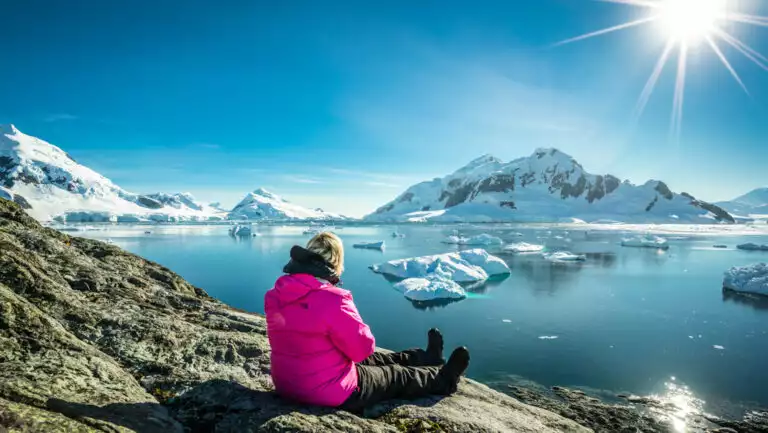 Woman in pink jacket sits on gray rock overlooking icy bay with glassy water & snowy peaks beyond on the Antarctic Peninsula.