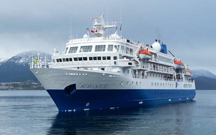 Blue & white Seaventure small expedition ship in Antarctica on an overcast day with calm seas & snowcapped mountains behind.