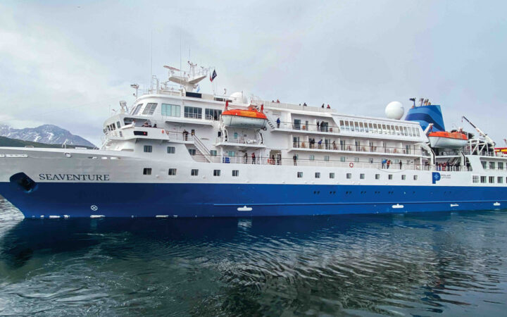 Exterior of Seaventure Antarctica expedition ship with blue hull & white upper decks with orange lifeboats, sitting in calm water.
