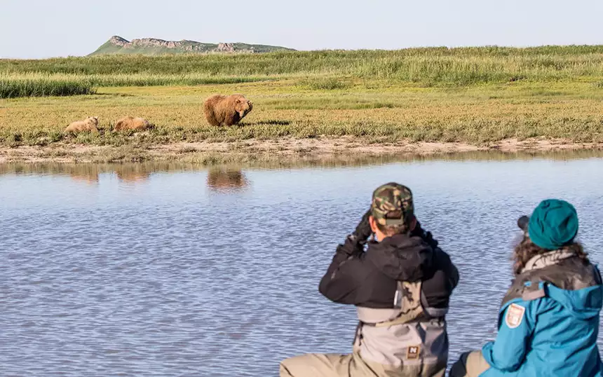 Two adventure travelers in a zodiac point binoculars towards a brown bear in a grass field with water in between them 