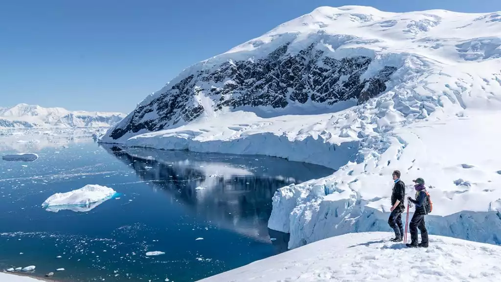 2 Antarctica air Cruise travelers stand in snowshoes atop a snowy peak overlooking a glassy bay with icebergs in the sun.
