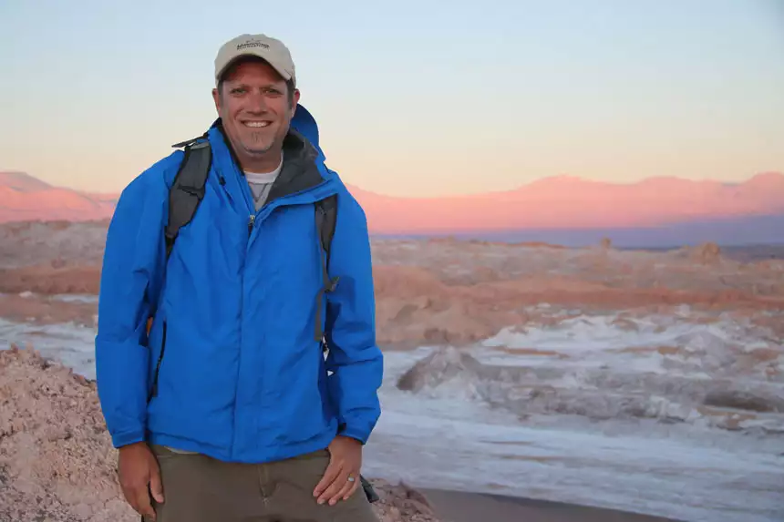 Man in blue jacket & tan hat stands atop a peak in the desert at sunset with pink & purple-hue mountains behind.