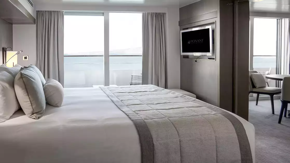 Deluxe Suite aboard L'Austral expedition ship, showing king bed, table, TV, chair & white-&-copper appointments.
