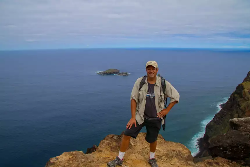 A traveler in a tan shirt and backpack stands on a cliff in Easter Island overlooking a small island in the ocean