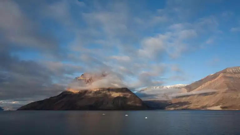 Clouds pass over jagged tan rock cliffs at top of peak overlooking calm fjord during an east & south Greenland Explorer cruise.