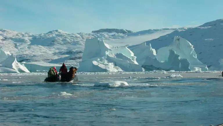 Zodiac boat with East & South Greenland travelers approaches a massive iceberg among a snowy landscape on a sunny day.