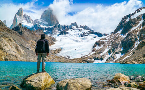 Explora El Chalten traveler stands on a boulder by a turquoise lake with a hanging snowfield under jagged peaks in the distance.