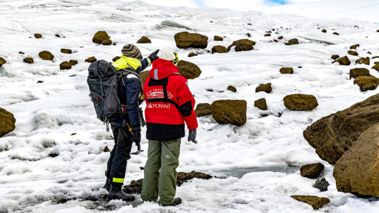 Guide with backpack & Antarctica traveler in red jacket stand atop snow & boulders field, looking around on a cloudy day.