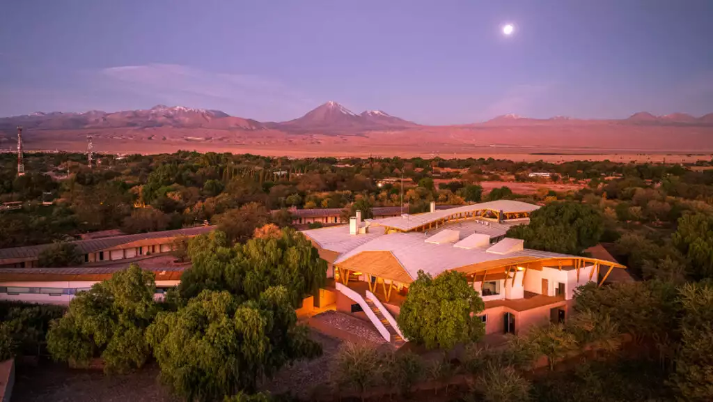 Aerial view of Explora Atacama Lodge in Chile at sunset, with white & wood construction among green trees & nearby desert.