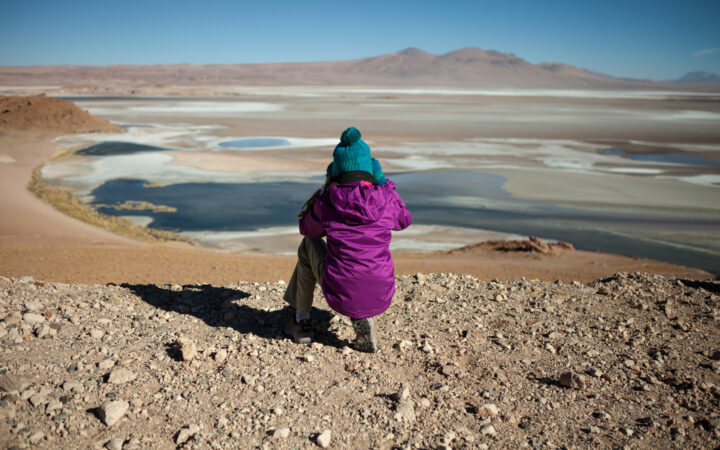 Solo desert hiker in purple coat & teal knit hat kneels at the summit to photograph the sandy red, beige & blue valley below.