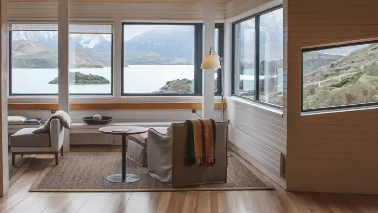 Living room at Explora Torres del Paine Lodge with beige padded chairs, small wood tables, lamps & walls of view windows.