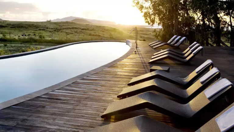 Long oval pool beside wood deck with modern black woven chaise lounge chairs at dusk, at Explora Rapa Nui lodge.