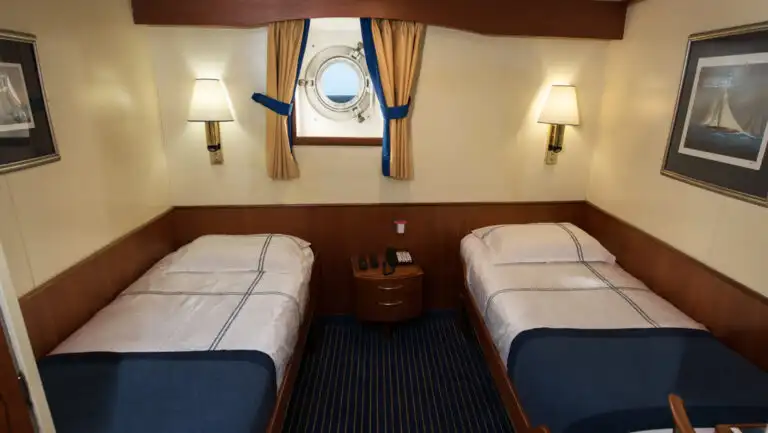 Lower Deck Twin cabin on G Expedition ship with 2 twin beds in blue & white linens, wood accents & 1 porthole.