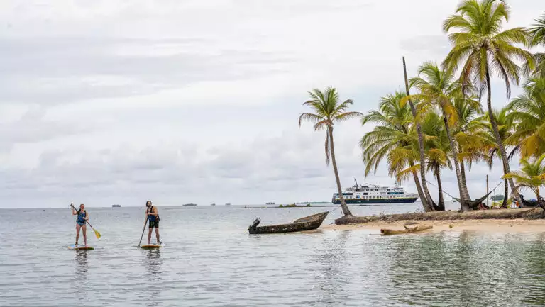 Man & woman paddle stand-up paddlebaords by shore of tiny island with white-sand beach & palm trees as ship sits nearby in Panama.