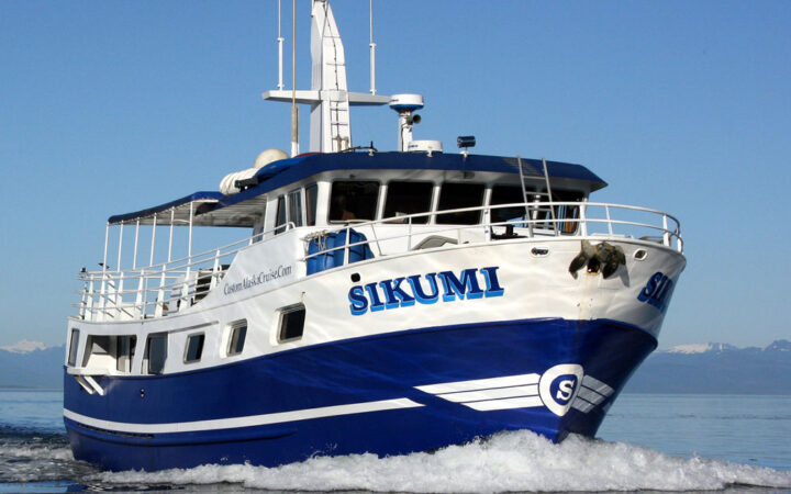 Sikumi small Alaska ship cruises in calm water on a sunny day, showing her dark blue & white exterior, main deck, wheelhouse & antennae.
