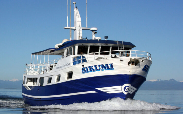 Sikumi small Alaska ship cruises in calm water on a sunny day, showing her dark blue & white exterior, main deck, wheelhouse & antennae.